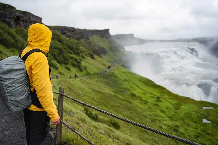 A person wearing a yellow rain jacket and carrying a backpack, standing on a path overlooking a misty waterfall and lush green landscape in Iceland during July.