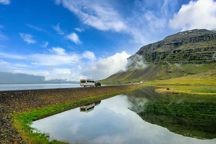 A camper van and car traveling along a road next to a serene lake in Iceland in July, with mountains and blue sky reflecting in the water.