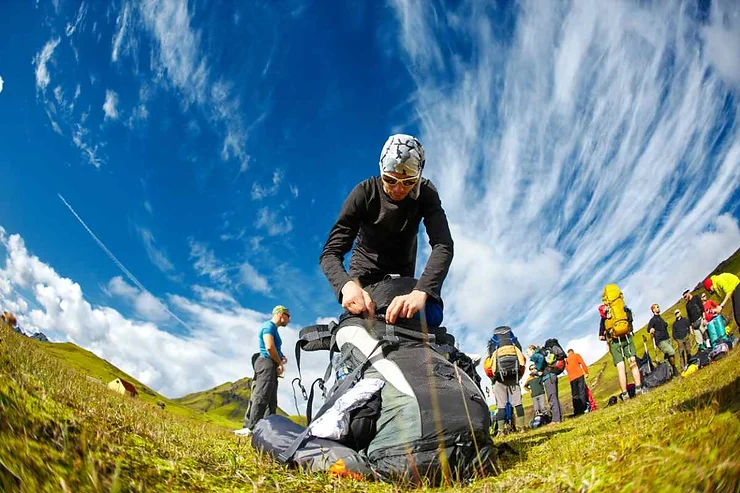 A group of people preparing for an adventure in Iceland in July, with a person in the foreground packing a large backpack under a vibrant blue sky with scattered clouds.