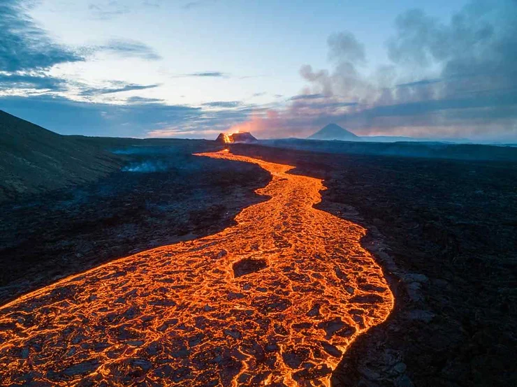 A striking view of a volcanic eruption in Reykjanes Peninsula, Iceland, showing a glowing river of lava flowing from the erupting volcano against a dramatic twilight sky.