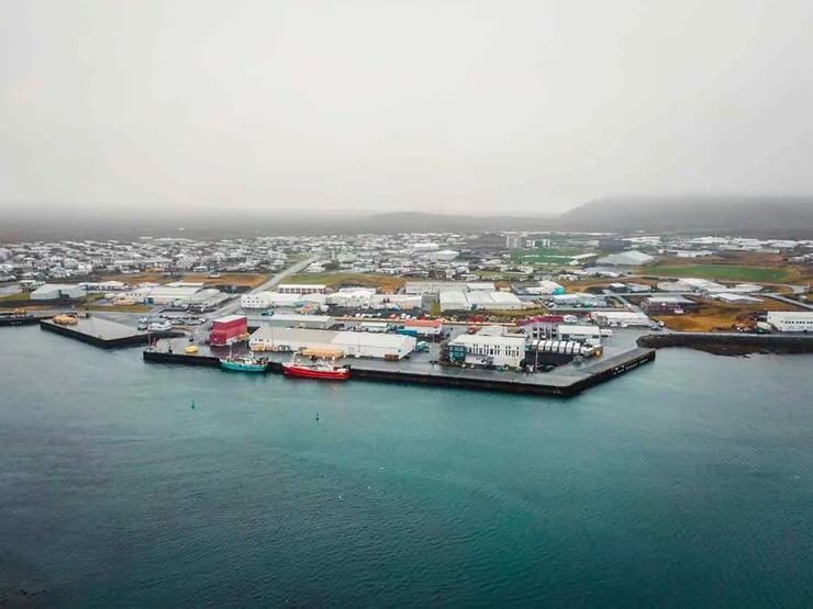 An aerial view of the coastal town of Grindavík in the Reykjanes Peninsula, Iceland, showcasing the harbor with colorful boats docked and a spread of buildings under a misty sky.