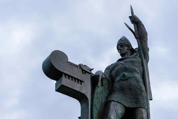 A close-up view of a historical Viking statue in Reykjanes Peninsula, Iceland, depicting a warrior in chainmail holding a spear and shield, with detailed carvings on the statue's base.