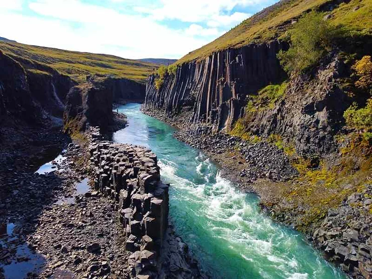 Aerial view of Studlagil Canyon in Iceland, showcasing the turquoise river winding through dramatic basalt column formations and lush green hillsides.