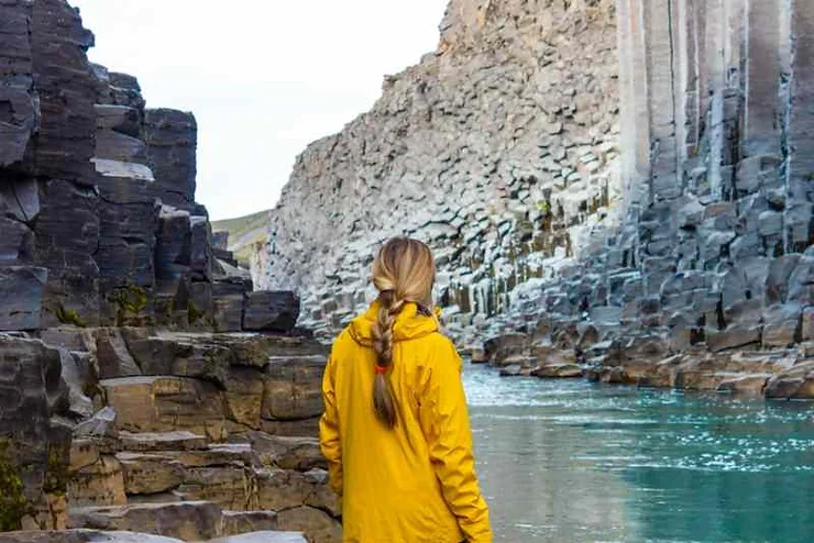 A hiker in a yellow jacket stands beside the turquoise river in Studlagil Canyon, Iceland, surrounded by towering basalt columns.