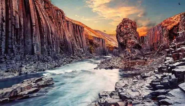 A stunning view of Studlagil Canyon in Iceland during sunset, with the sun casting a warm glow on the basalt columns and a serene river flowing through the canyon.