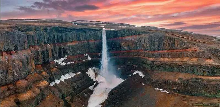 A breathtaking view of a waterfall cascading down the cliffs of Studlagil Canyon in Iceland during sunset, with the sky painted in shades of pink and orange.