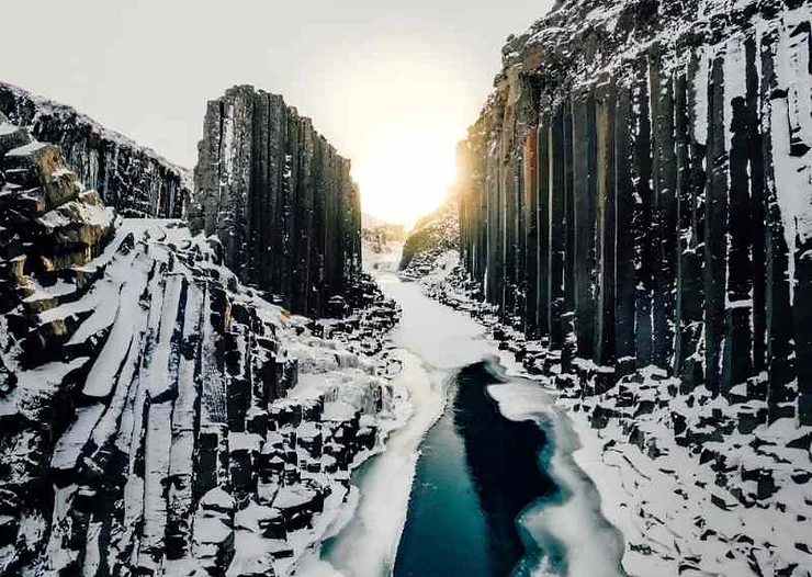 A breathtaking winter scene in Studlagil Canyon, Iceland, with snow-covered basalt columns and a partially frozen turquoise river under the bright sunlight.