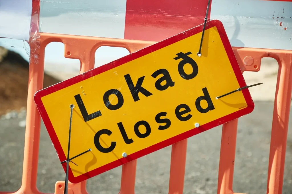 A traffic sign in Iceland with the Icelandic word 'loka' and its English translation 'closed' displayed on a yellow background with red border. The sign is attached to an orange-white barricade.