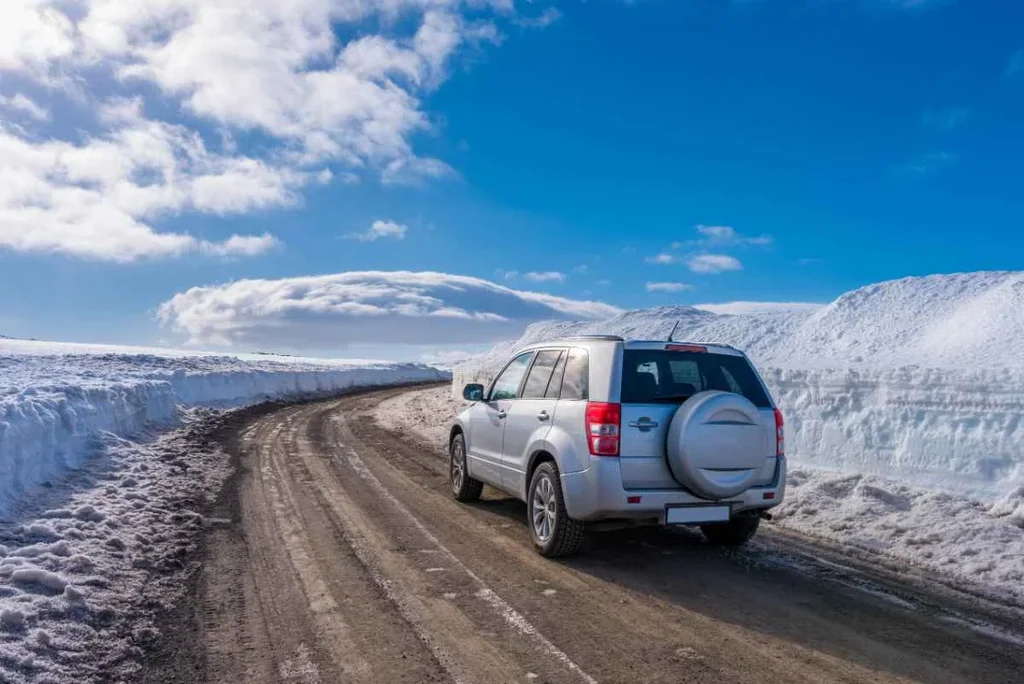 A white SUV driving on a snow-cleared road surrounded by high snowbanks under a bright blue sky in Iceland.