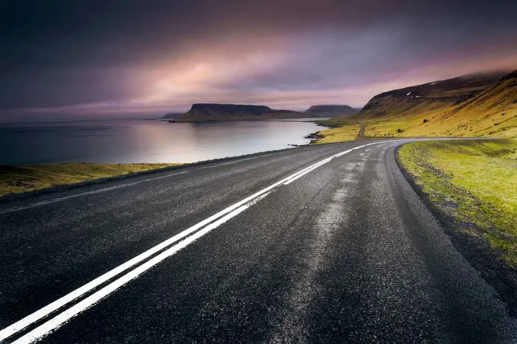 A scenic Icelandic coastal highway at dusk, with a dramatic sky reflecting hues of purple and pink over the calm waters.