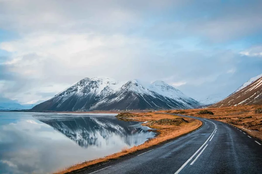 A scenic paved road by a calm lake with snow-capped mountains reflected on the water under a pastel sky.