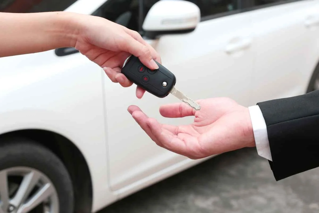 A person handing over a car key to another, with a white rental car in the background.