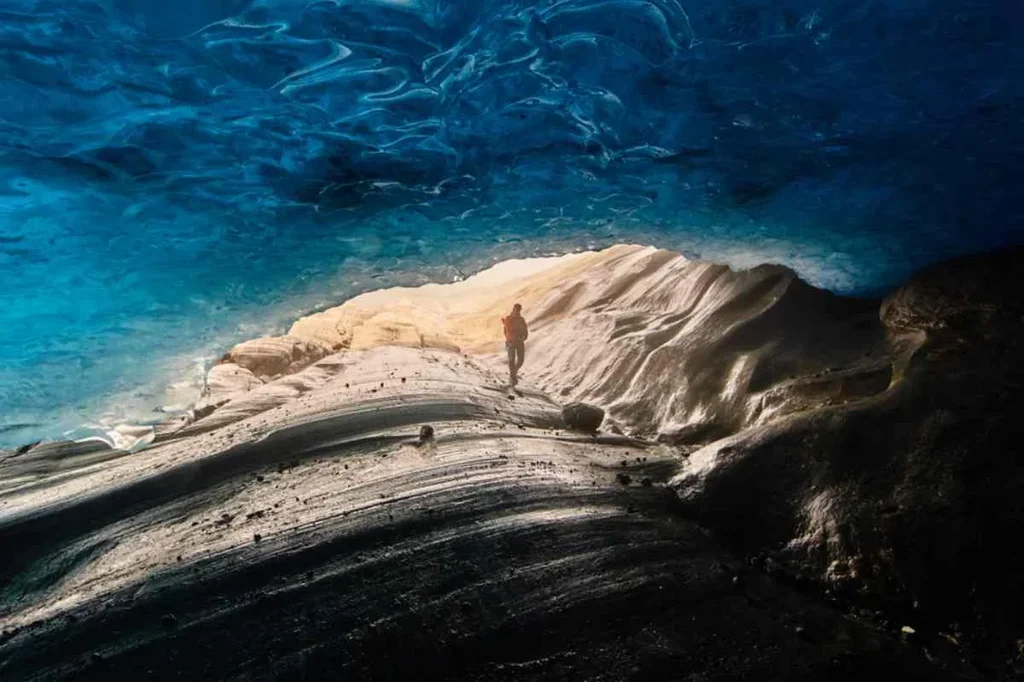 Person walking inside an ice cave with blue ice ceiling and rugged terrain, highlighting the natural beauty of Langjokull Glacier in Iceland.