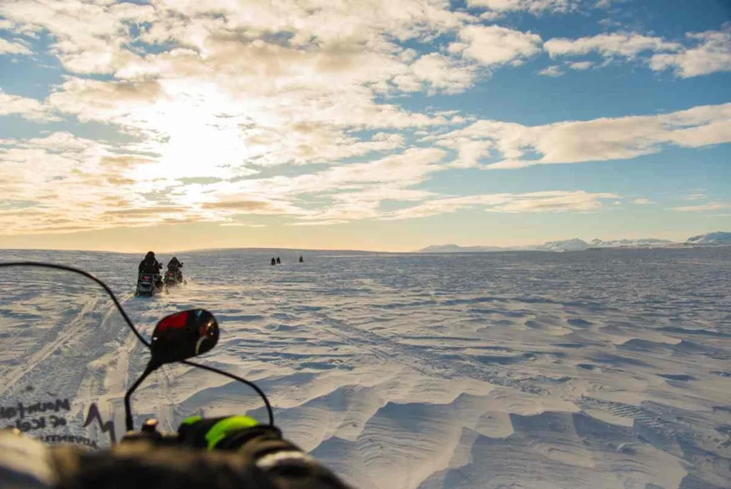 View from a snowmobile on Langjokull Glacier, with several snowmobilers in the distance under a partly cloudy sky at sunset, creating a dramatic and adventurous atmosphere.