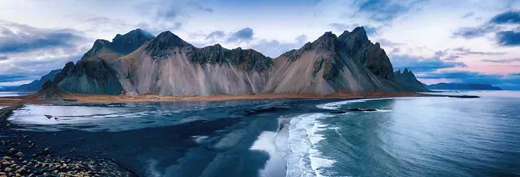 A panoramic view of Vestrahorn Mountain in Iceland, showcasing its rugged peaks and the black sand beach meeting the serene waters of the Atlantic Ocean at sunset.