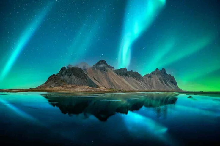 The Northern Lights dancing over Vestrahorn Mountain in Iceland, reflecting beautifully on the calm waters below, with vibrant green and blue hues lighting up the night sky.