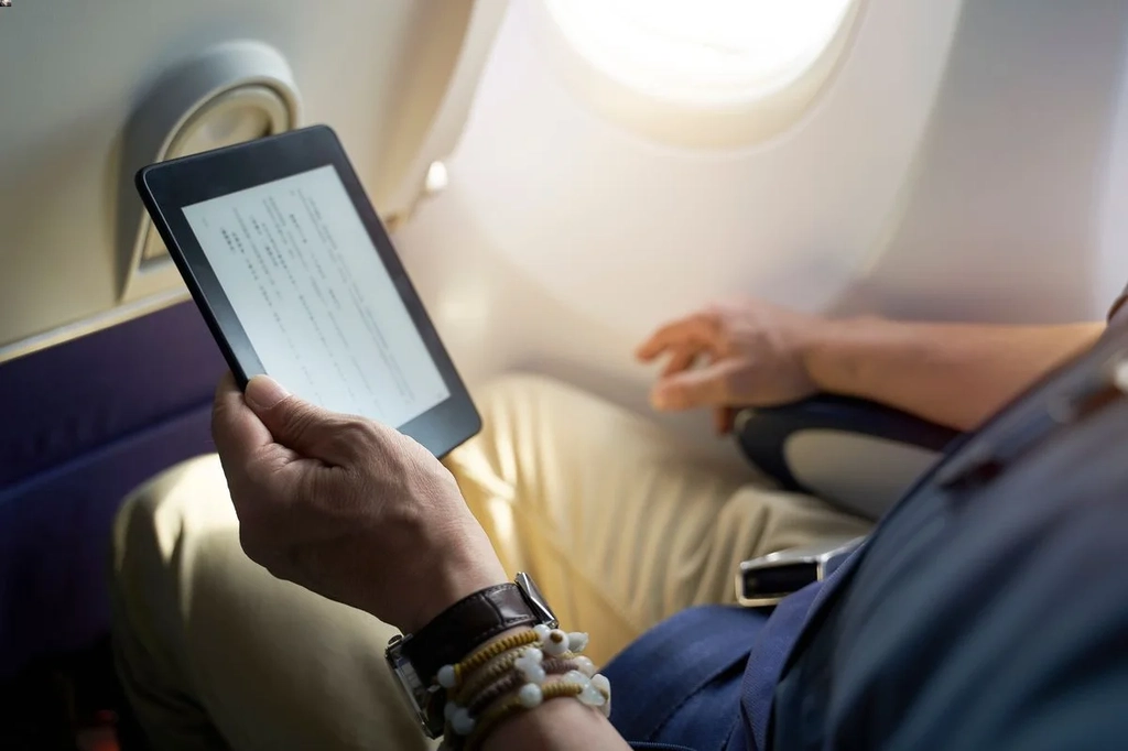 A person reading an e-book on a tablet while sitting in an airplane, with a window view in the background, highlighting a comfortable and modern travel experience.