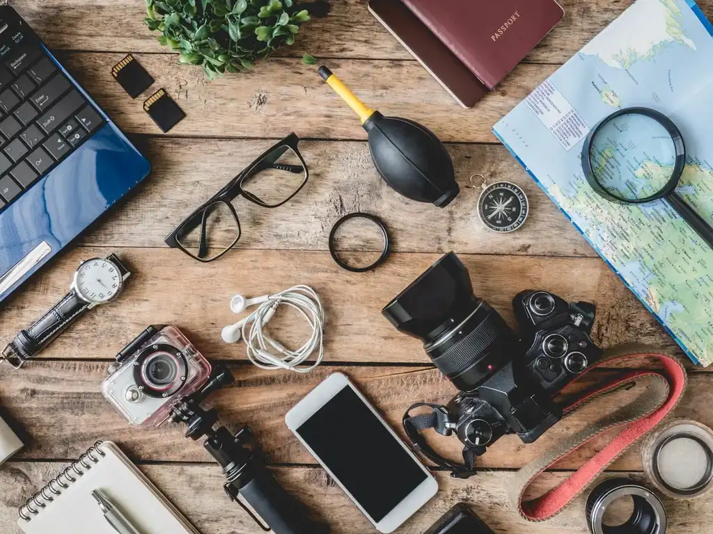 A flat lay of travel essentials on a wooden table, including a laptop, camera, smartphone, headphones, watch, glasses, map, passport, compass, magnifying glass, and memory cards, showcasing the necessary gadgets and items for a trip.