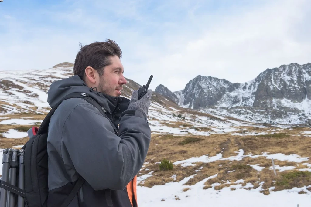 A man in a winter jacket using a walkie-talkie while standing in a snow-covered mountainous area, emphasizing outdoor communication and safety during hiking or trekking.