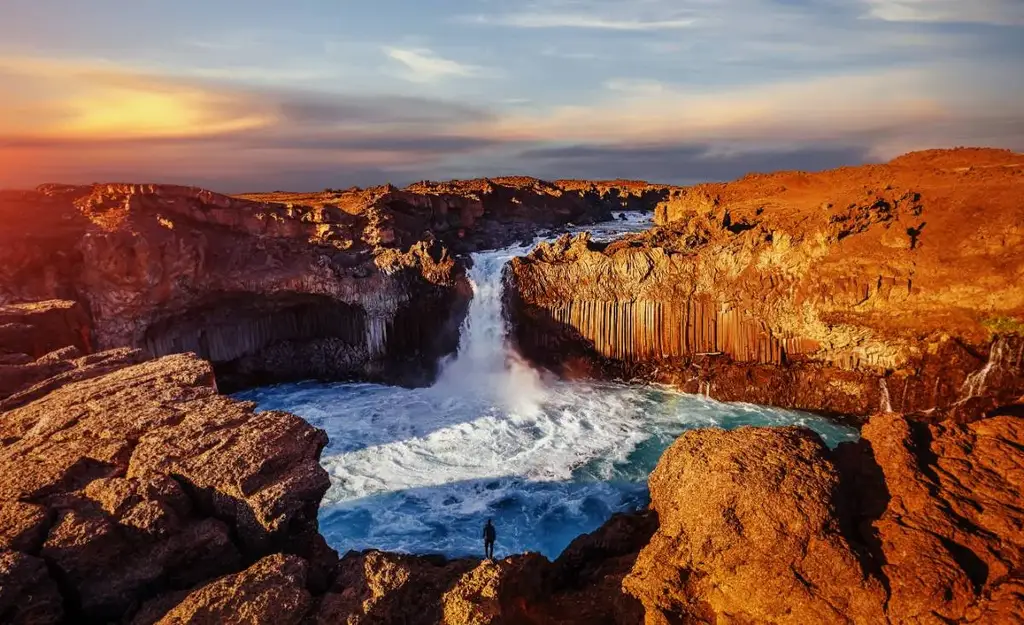 Aldeyjarfoss waterfall in Iceland during sunset, with cascading water flowing into a turbulent pool surrounded by rugged basalt columns and red-tinged rocky terrain.