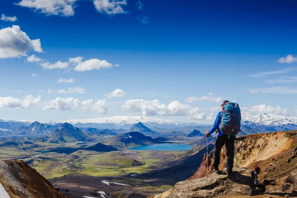 A hiker with a large backpack and hiking poles stands on a rocky outcrop, overlooking the stunning, expansive landscape of Iceland. The view includes rolling green hills, distant mountains, snow-capped peaks, and a serene blue lake under a clear, bright blue sky with scattered clouds.