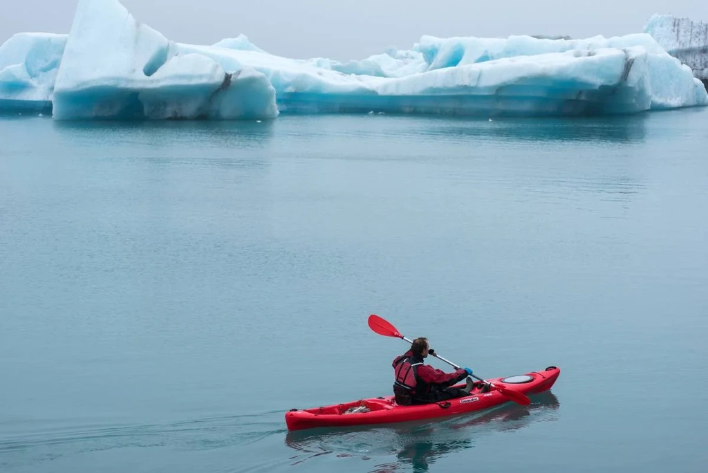 A person wearing a red jacket paddles a red kayak on a calm glacier lagoon, surrounded by large, blue-tinted icebergs. The scene is serene and peaceful, with the icy formations reflecting on the still water.