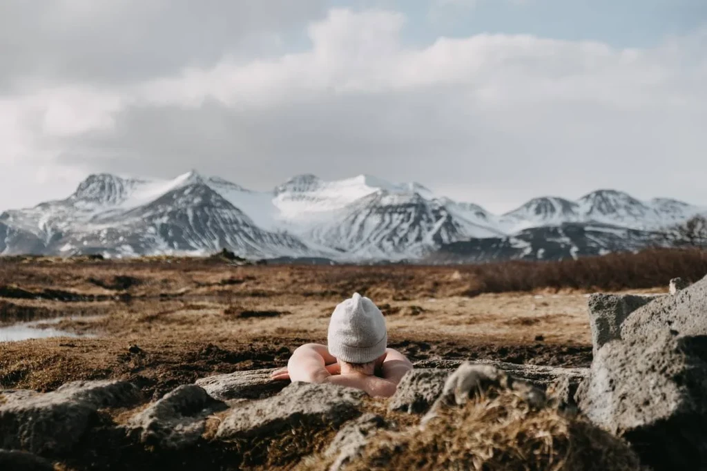 A person wearing a beanie relaxing in the Landbrotalaug hot spring in Iceland, surrounded by rocky terrain and overlooking snow-capped mountains under a cloudy sky.