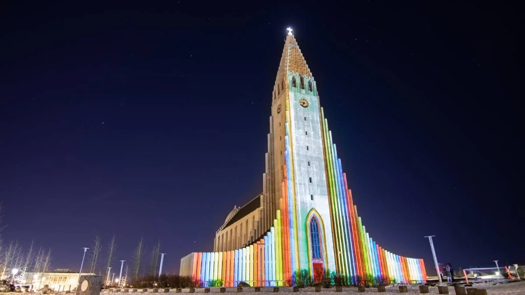 Hallgrímskirkja, the iconic church in Reykjavik, illuminated with vibrant rainbow colors at night. The tall, striking structure stands against a dark sky, with the colorful lights emphasizing its unique architectural design and creating a stunning visual spectacle.