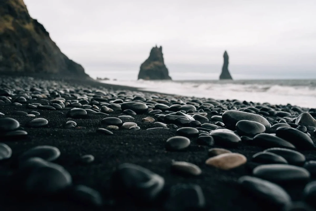 A close-up of smooth, dark pebbles on the black sand beach of Reynisfjara in Iceland. The beach stretches out to the sea with towering basalt sea stacks rising from the water in the background under a cloudy sky.