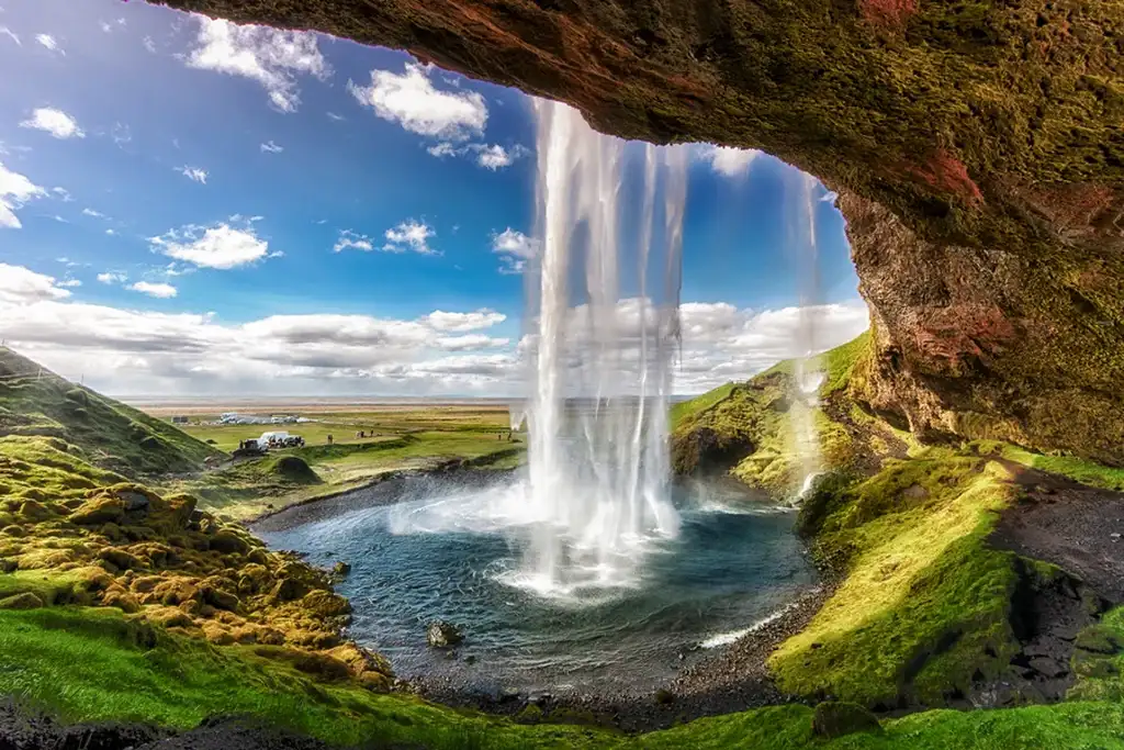 View from behind Seljalandsfoss waterfall in Iceland, with water cascading down in front of a lush green landscape and a bright blue sky dotted with white clouds.