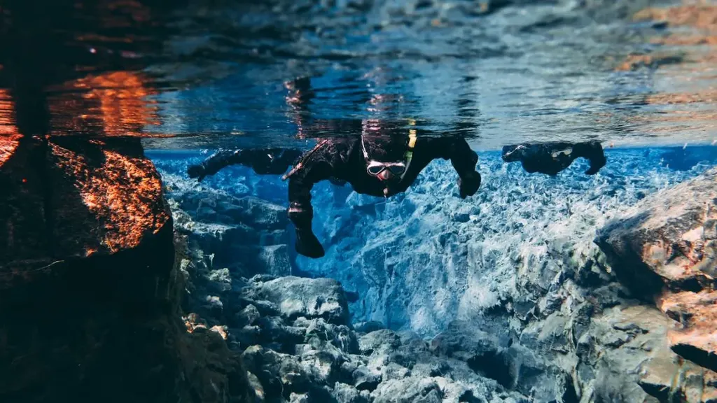 A person wearing a wetsuit and snorkel gear swims in the crystal-clear waters of the Silfra Fissure. The underwater scene reveals a stunning rocky landscape with vibrant blue and orange hues, showcasing the unique geological formations of the fissure.