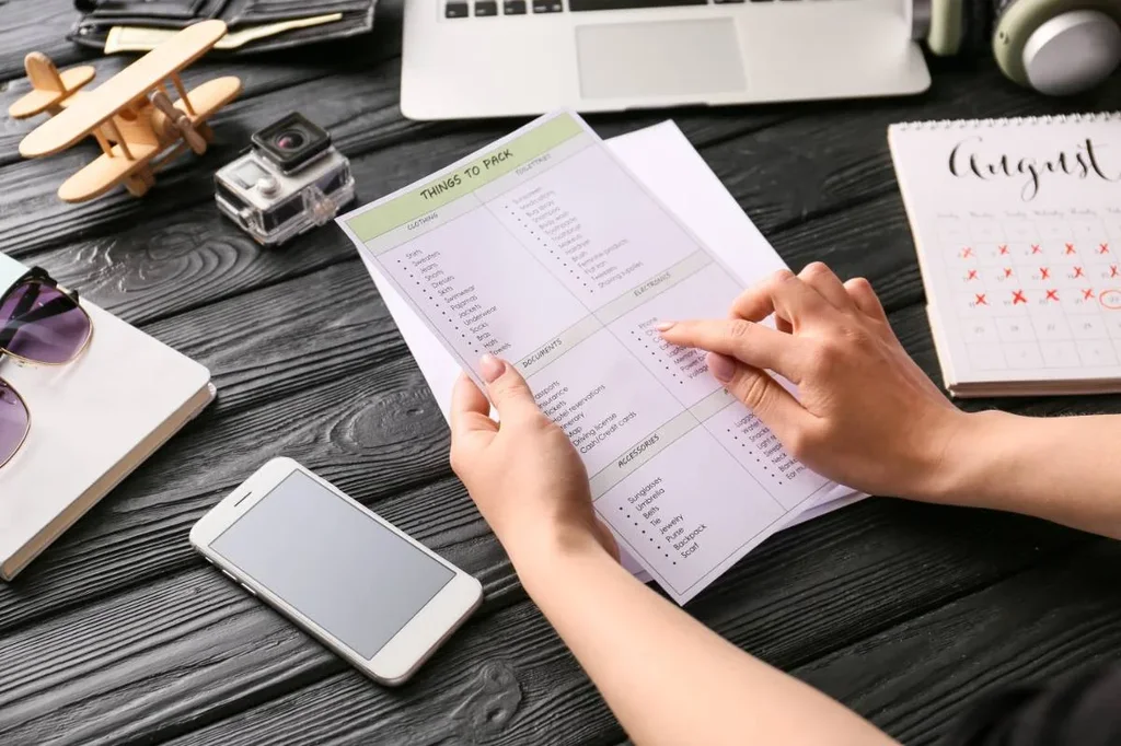 A person reviewing a things to pack checklist for a trip surrounded by travel items like phone, sunglasses, GoPro camera, small plane model, and calendar marked August