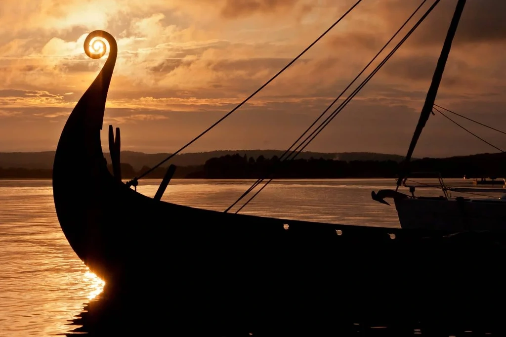 Silhouette of a Viking ship with a prominent curved prow against the backdrop of a serene sunset. The golden light reflects on the calm water, highlighting the ship's intricate design and evoking a sense of historical adventure and exploration.