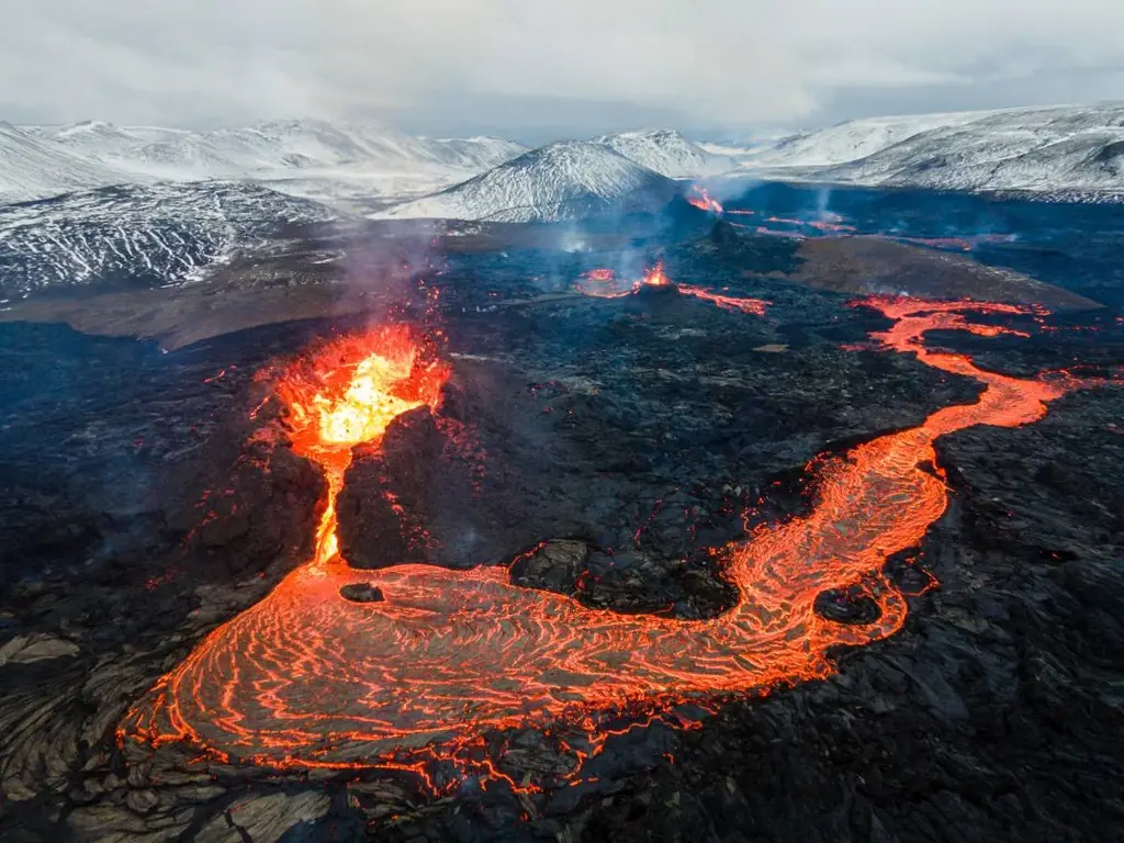 Aerial view of a volcanic eruption in Iceland, with bright red lava flowing from the crater, creating a molten river on the dark, rugged terrain. Snow-covered mountains can be seen in the background under a cloudy sky.