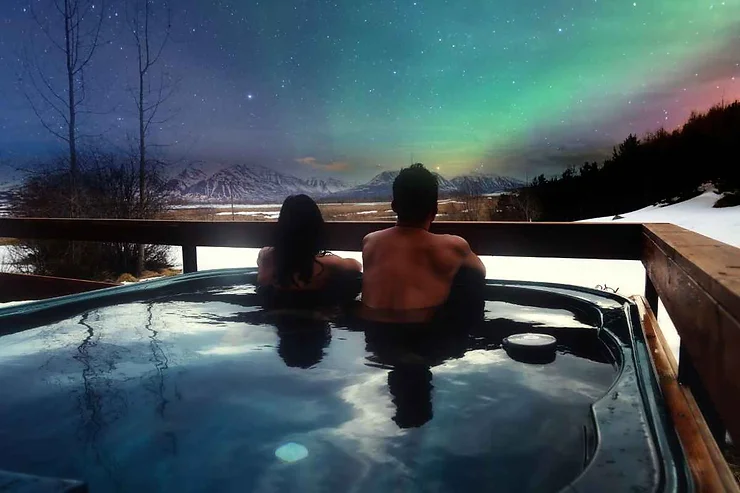 A couple relaxing in a hot tub with a view of the Northern Lights and snow-covered mountains at a hotel along the Outer Ring Road in Iceland.