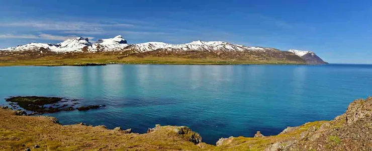 A panoramic view of Borgarfjordur fjord in Iceland, showcasing snow-capped mountains, clear blue water, and rugged terrain under a clear sky.