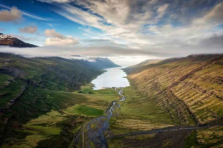 A breathtaking aerial view of Mjoifjordur fjord in Iceland, featuring a winding river, lush green valleys, steep mountains, and a partly cloudy sky.