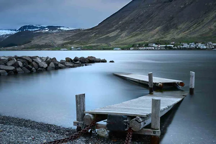 A serene view of Patreksfjordur fjord in Iceland, featuring a small wooden pier extending into calm waters, with rocky shores and mountainous terrain in the background.