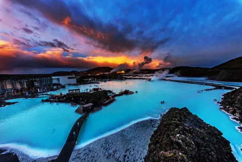 The Blue Lagoon in Iceland at sunset, showcasing its stunning geothermal waters. The lagoon's milky blue water contrasts beautifully with the dark volcanic rocks surrounding it. The sky is painted with vibrant hues of orange, pink, and purple as the sun sets, casting a warm glow over the scene. Steam rises from the hot springs, adding to the mystical atmosphere. The facility's modern buildings and walkways are visible in the background, along with a few bathers enjoying the soothing waters. This image captures the serene and otherworldly beauty of one of Iceland's most famous natural attractions.