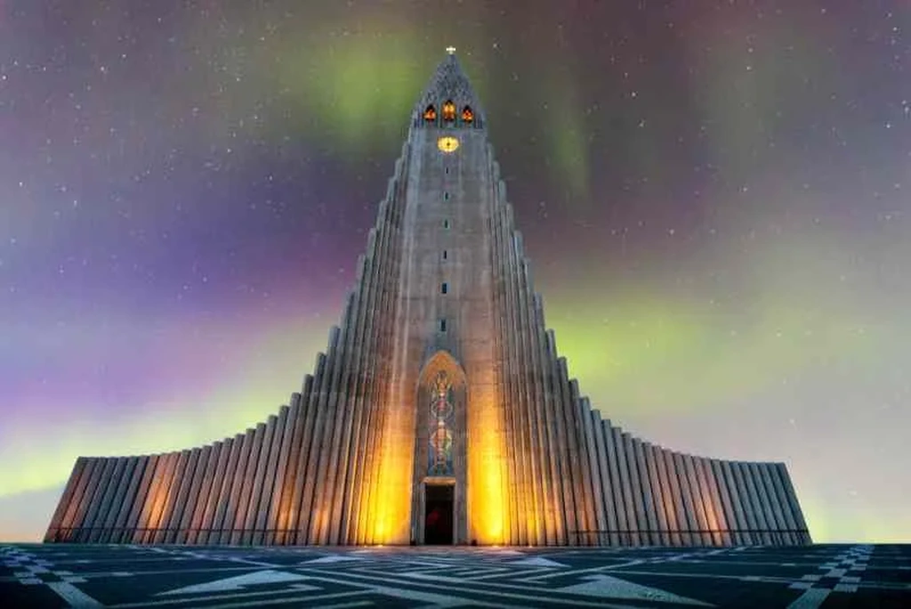 Hallgrímskirkja, the iconic church in Reykjavik, lit up at night with warm golden lights highlighting its distinctive vertical lines. The sky above is illuminated with a beautiful display of the Northern Lights, creating a mesmerizing backdrop for the stunning architecture of the cathedral.