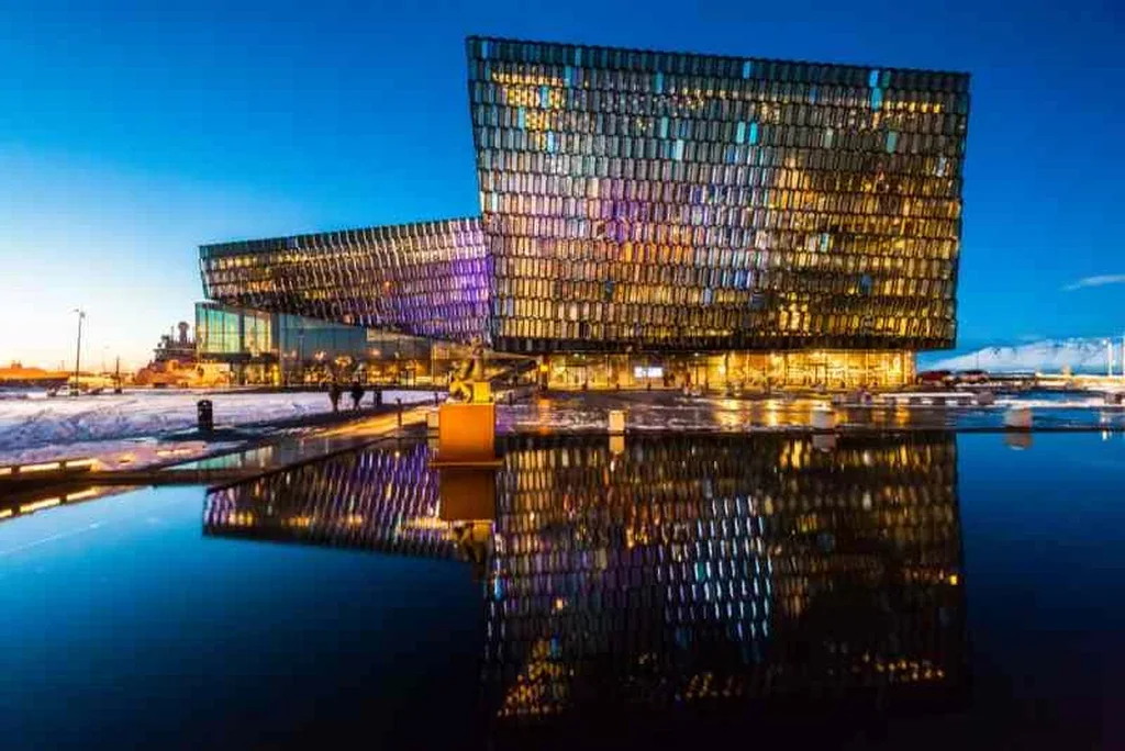 Harpa Concert Hall in Reykjavik, Iceland, illuminated at night. The modern glass structure reflects an array of colorful lights, creating a vibrant display. The building's unique geometric design features thousands of glass panels that shimmer in various shades, including purple, blue, and gold. In front of the concert hall, a calm body of water mirrors the stunning architecture, enhancing the visual effect. The surrounding area is softly lit, with a few people walking by, adding a sense of scale to the scene. The clear, deep blue sky provides a perfect backdrop, highlighting the architectural marvel of Harpa against the evening landscape.