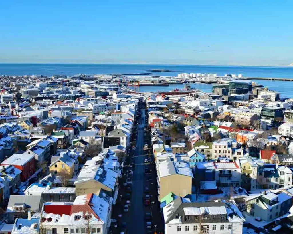 Aerial view of Reykjavik, Iceland, showcasing Laugavegur Street stretching straight through the city. The buildings, painted in a variety of pastel colors, are dusted with snow, adding to the wintry charm. The North Atlantic Ocean provides a stunning blue backdrop to the urban landscape. In the distance, industrial buildings and storage tanks are visible near the waterfront. The image captures the vibrant and bustling essence of Reykjavik under a clear blue sky, with the contrast of snowy rooftops and colorful architecture.