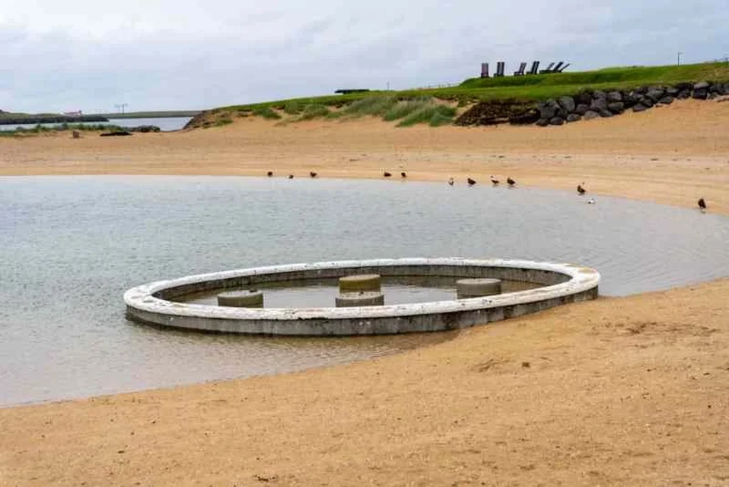 A serene view of Nauthólsvík Geothermal Beach in Iceland. The image features a circular concrete structure partially submerged in the shallow waters of the beach. The sandy shore is lined with small birds, and grassy dunes rise gently in the background. The scene is calm and inviting, with overcast skies adding to the tranquil atmosphere.