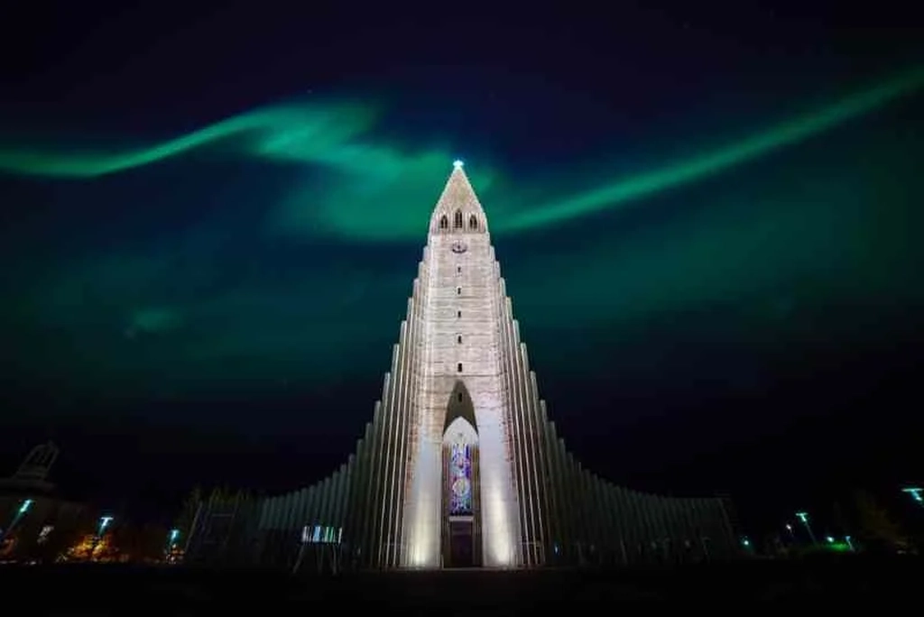 Hallgrímskirkja, the landmark church in Reykjavik, beautifully illuminated at night with a white glow that accentuates its unique architectural lines. Above the church, the Northern Lights paint the sky with vibrant green hues, creating a stunning and serene atmosphere over the Icelandic capital.