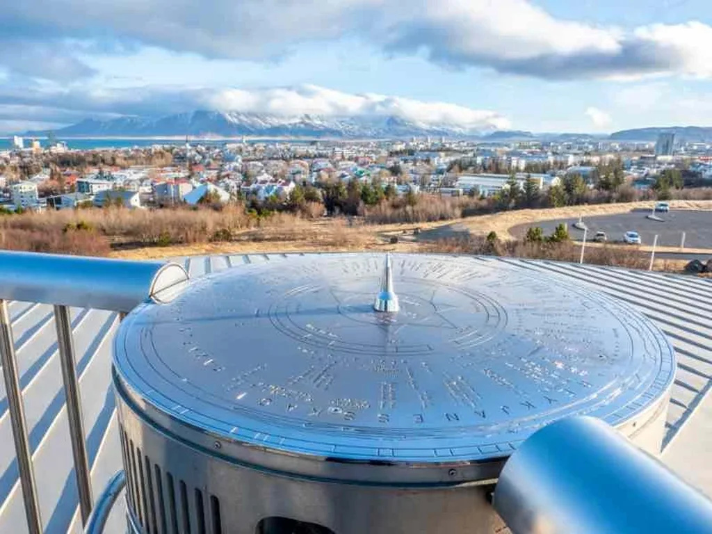 A panoramic view from the Perlan Observatory in Reykjavik, Iceland. In the foreground, a metallic dial points towards various landmarks, with engraved names and directions. The city of Reykjavik spreads out below, showcasing a mix of modern buildings and traditional houses. In the distance, snow-capped mountains are partially covered by clouds, while the blue waters of the surrounding bay reflect the sky. The image captures the clear and bright atmosphere of a sunny day, highlighting the stunning landscape and architectural beauty of Iceland's capital.