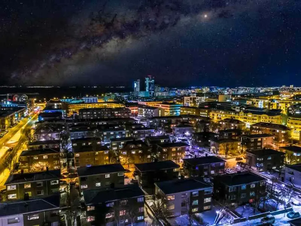 A vibrant aerial view of Reykjavik at night, showcasing the city's lively nightlife. The buildings are illuminated with warm lights, creating a contrast against the dark sky. In the background, the cityscape features taller buildings and twinkling lights, while the Milky Way stretches across the sky, adding a touch of cosmic beauty to the urban scene.