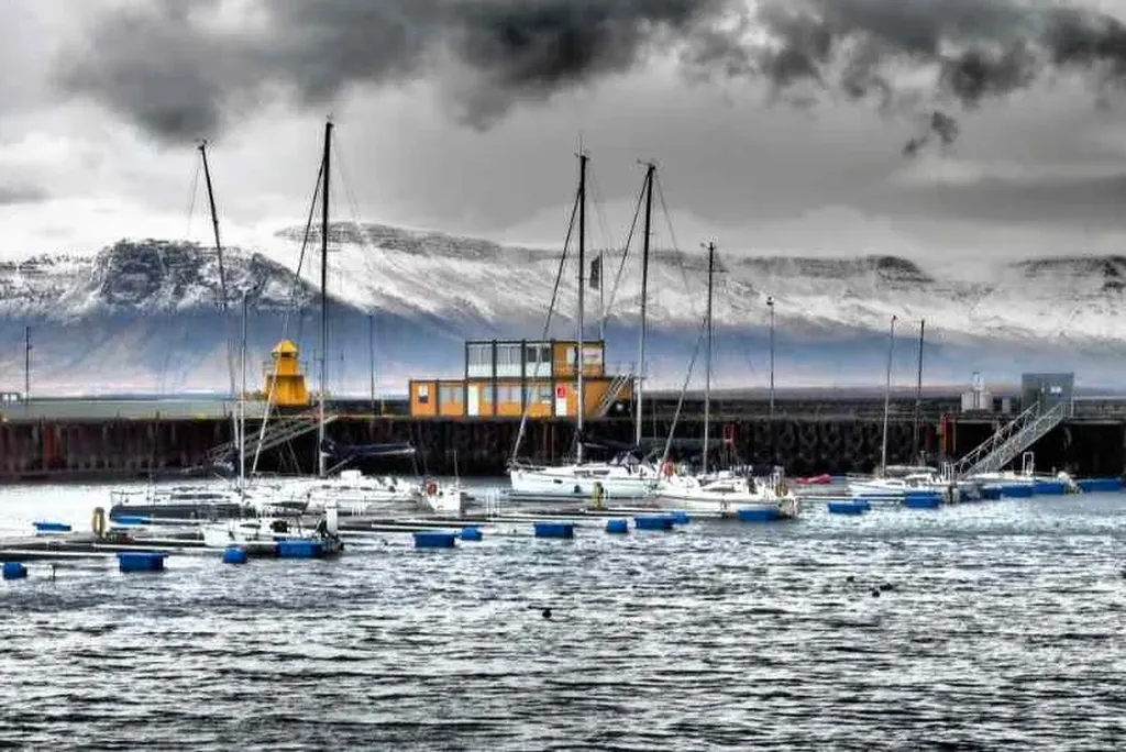 Reykjavik Old Harbour in Iceland, showcasing a collection of sailboats and yachts docked at the marina. The boats, with their tall masts, are neatly arranged along the wooden piers. In the background, a small yellow lighthouse and a modern building with large windows add a pop of color to the scene. The harbor is surrounded by a concrete seawall, providing protection from the sea. Behind the harbor, snow-capped mountains rise majestically, their peaks shrouded in a layer of dark, dramatic clouds. The contrast between the calm waters of the harbor and the rugged, wintry landscape creates a striking visual. The overcast sky enhances the moody atmosphere, highlighting the serene yet powerful nature of Iceland's coastal environment.