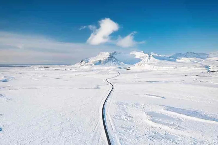 A long, winding road stretches through a vast, snow-covered landscape in Iceland during March. The surrounding area is blanketed in white, with distant mountains under a clear blue sky, creating a serene and picturesque winter scene.