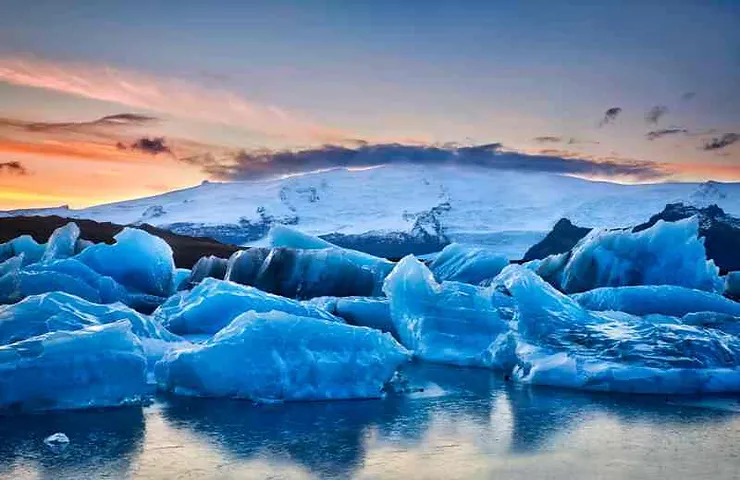 A stunning view of blue glaciers floating in a calm lagoon with a snow-covered mountain in the background. The sky is painted with hues of orange and pink as the sun sets, reflecting on the icy waters.