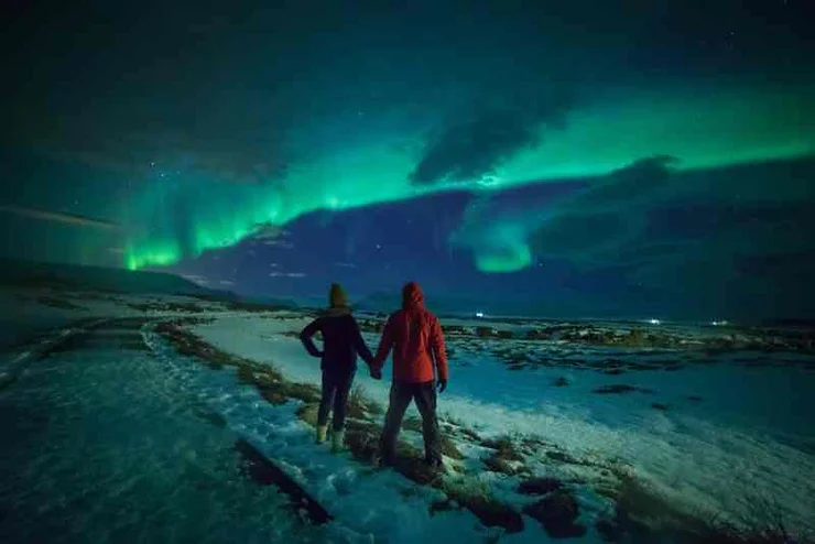 A breathtaking scene of the Northern Lights illuminating the night sky in Iceland, with two people holding hands, standing on a snow-covered path, marveling at the vibrant green and blue auroras above them.